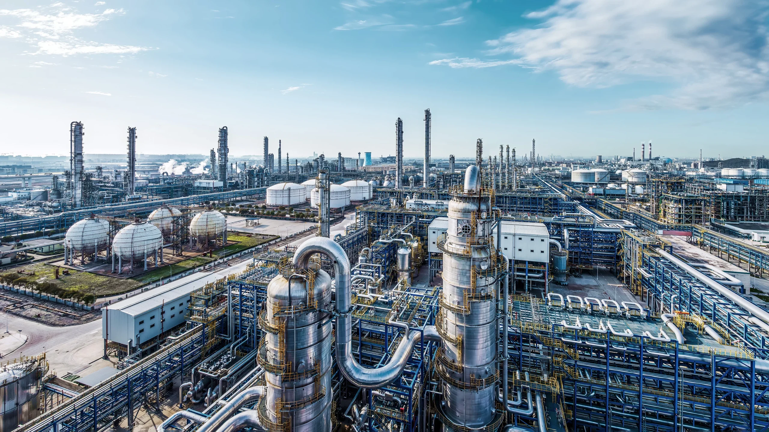 Sprawling oil refinery complex with tall distillation columns, spherical storage tanks, and a maze of interconnected pipes under a clear blue sky.